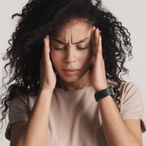 Pessoa com expressão de dor ou estresse, olhos fechados e mãos na cabeça. Veste camisa clara e está em ambiente neutro.