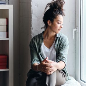 Pessoa sentada perto da janela, olhando para fora, em momento tranquilo. Usa camisa verde, calça marrom e tem cabelo preso. Ambiente interno com estante e parede branca.