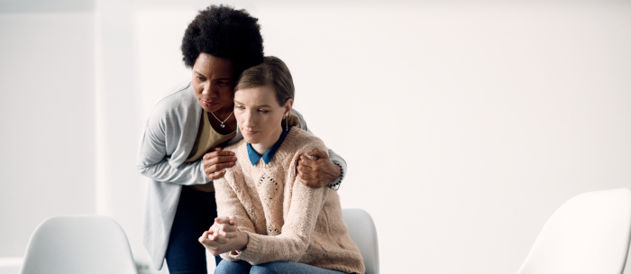 Duas pessoas em sala clara: uma sentada pensativa, outra em pé ao lado oferecendo apoio com o braço.